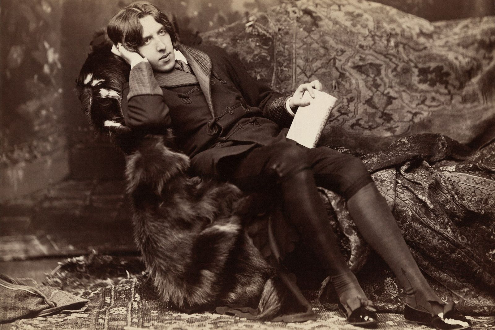 Oscar Wilde reclining in a fur-trimmed coat on a patterned couch, one hand to his head and a book in the other.