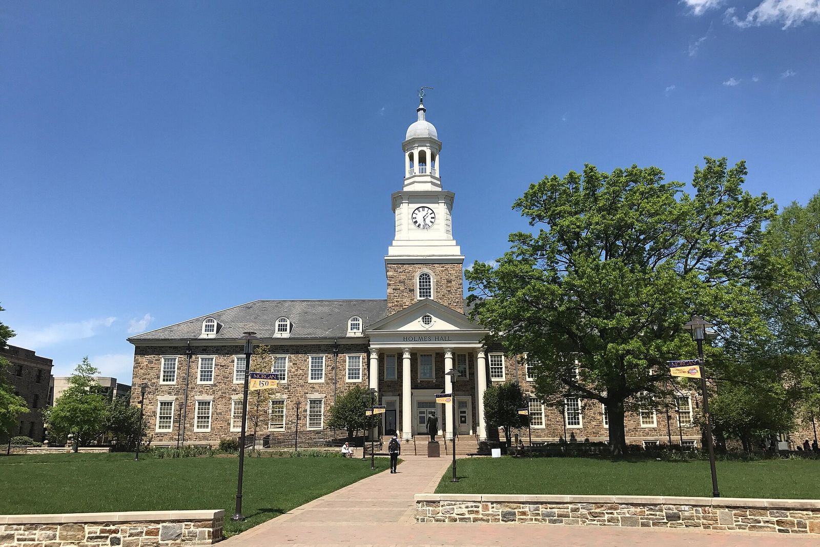 Stone Holmes Hall with a white clock tower stands behind a lawn and brick walkway on the Morgan State campus.