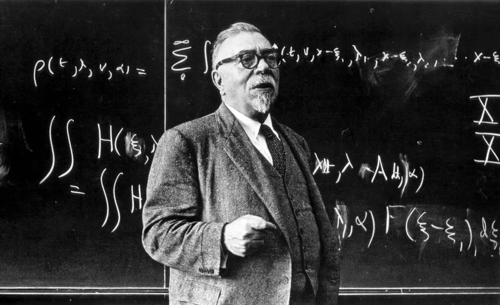 Norbert Wiener in glasses and a tweed jacket stands lecturing in front of a blackboard covered in equations.