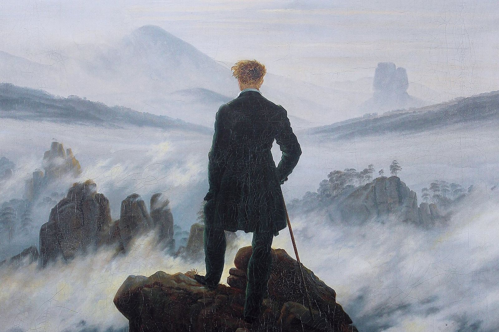 A man in a dark coat seen from behind stands on a rocky peak above a sea of fog and distant mountains.