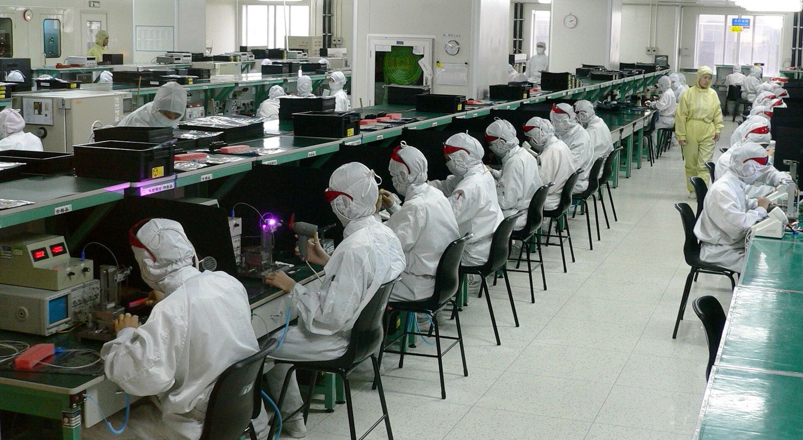 Rows of workers in white cleanroom suits and masks assembling electronics along factory benches.