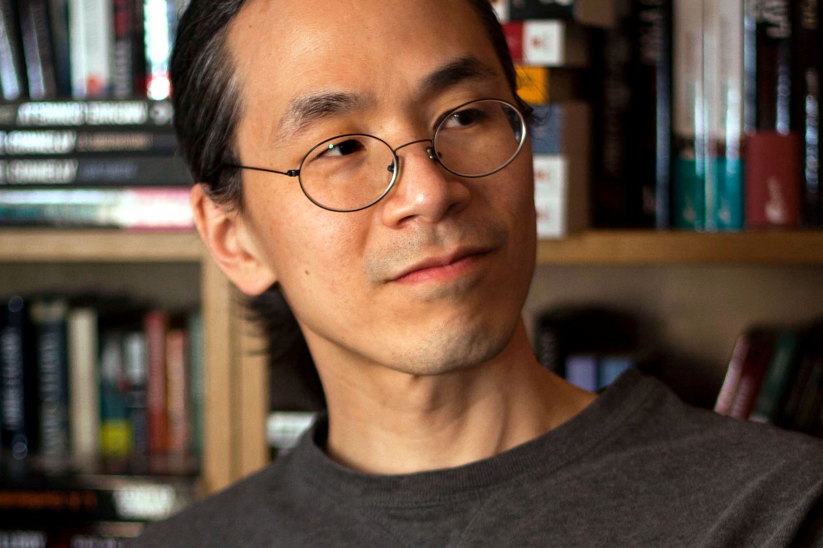 Writer Ted Chiang in glasses, seated in front of loaded bookshelves.