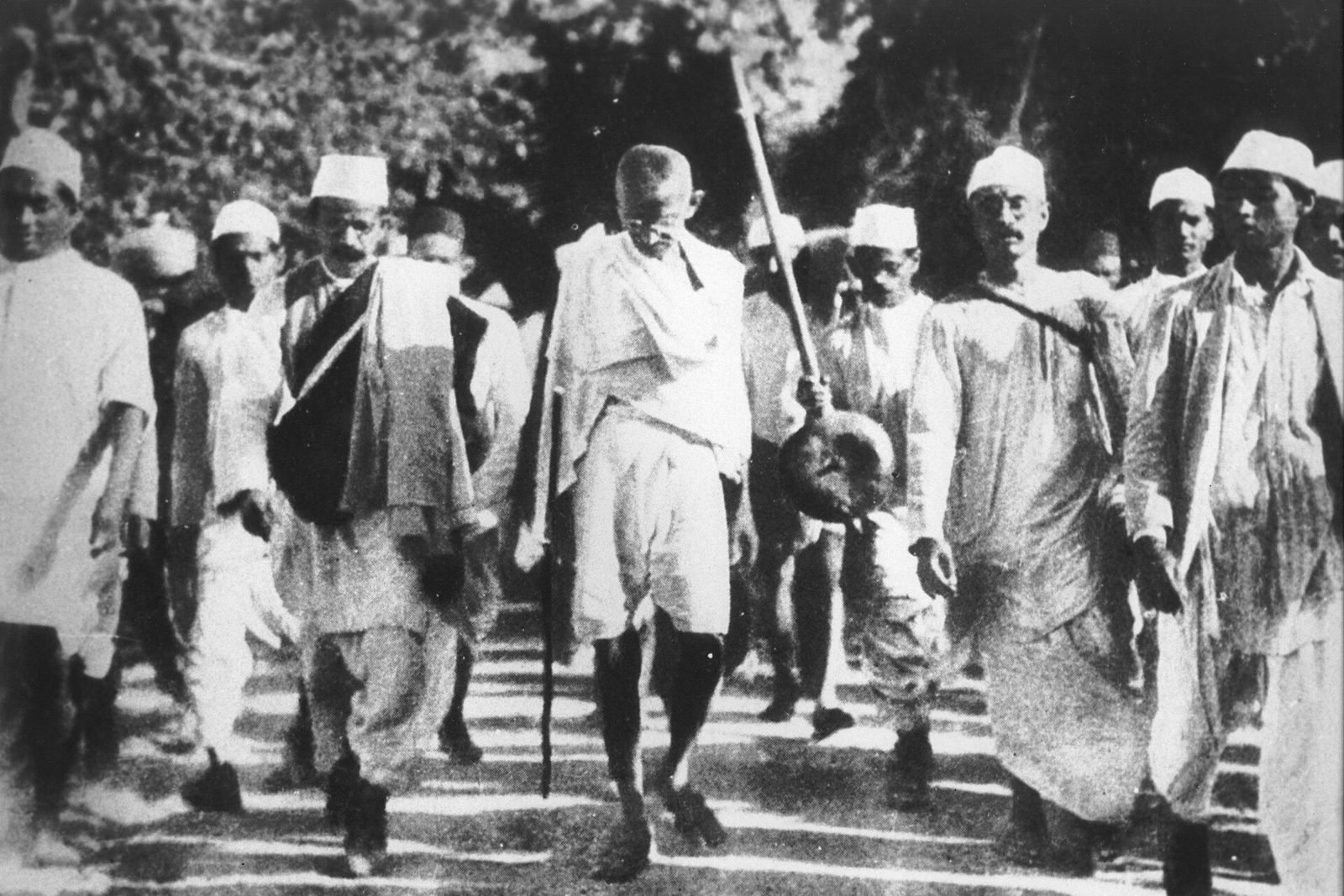 Gandhi walking at the head of a column of marchers on a tree-lined road.