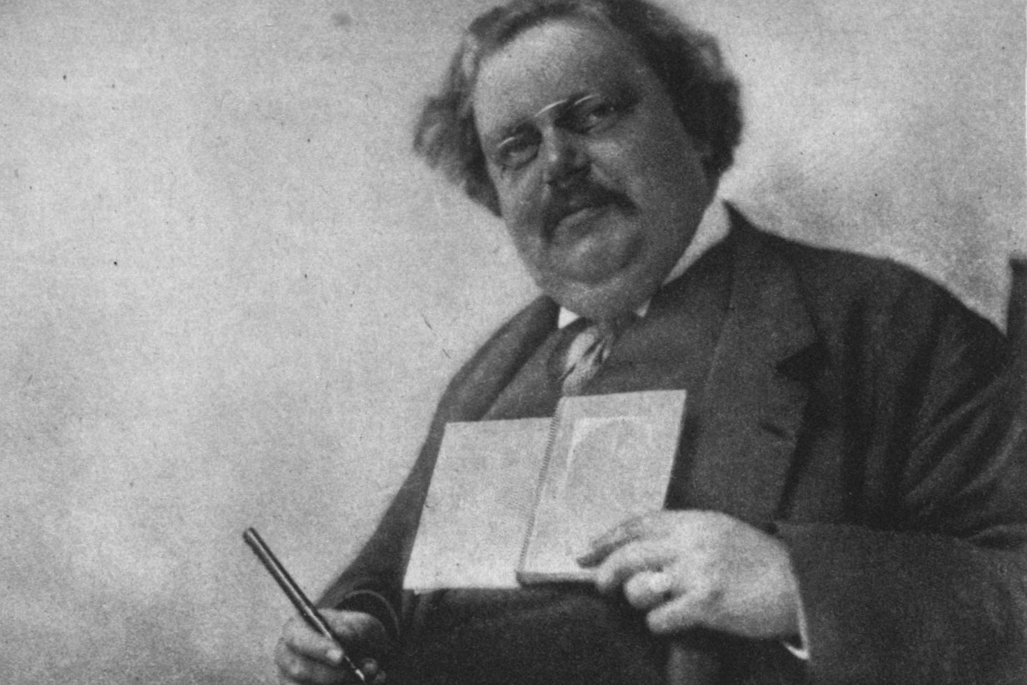 G. K. Chesterton, a heavyset man with a moustache and wavy hair, holds a pen and an open book.