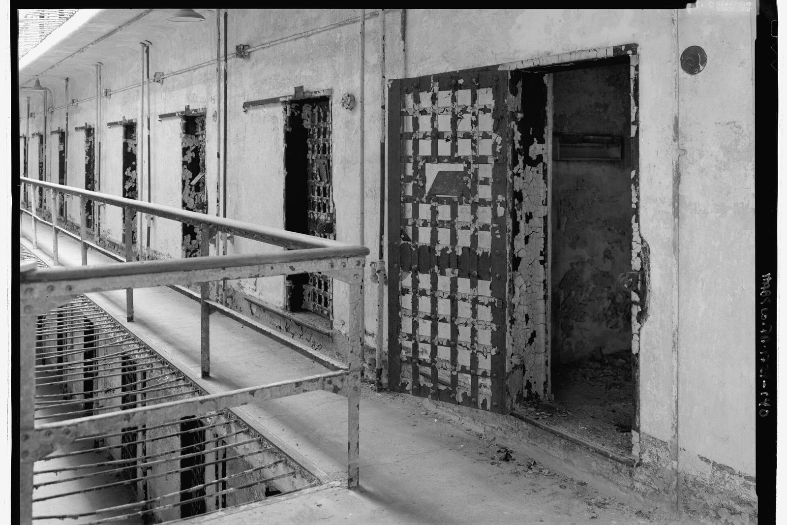 Black-and-white view along a prison tier with a heavy barred cell door standing open beside a railing.