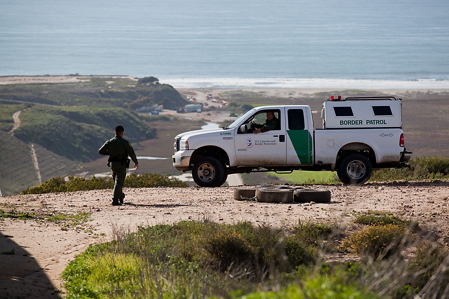 A Border Patrol agent on foot beside a Border Patrol truck on a hillside above the coast.