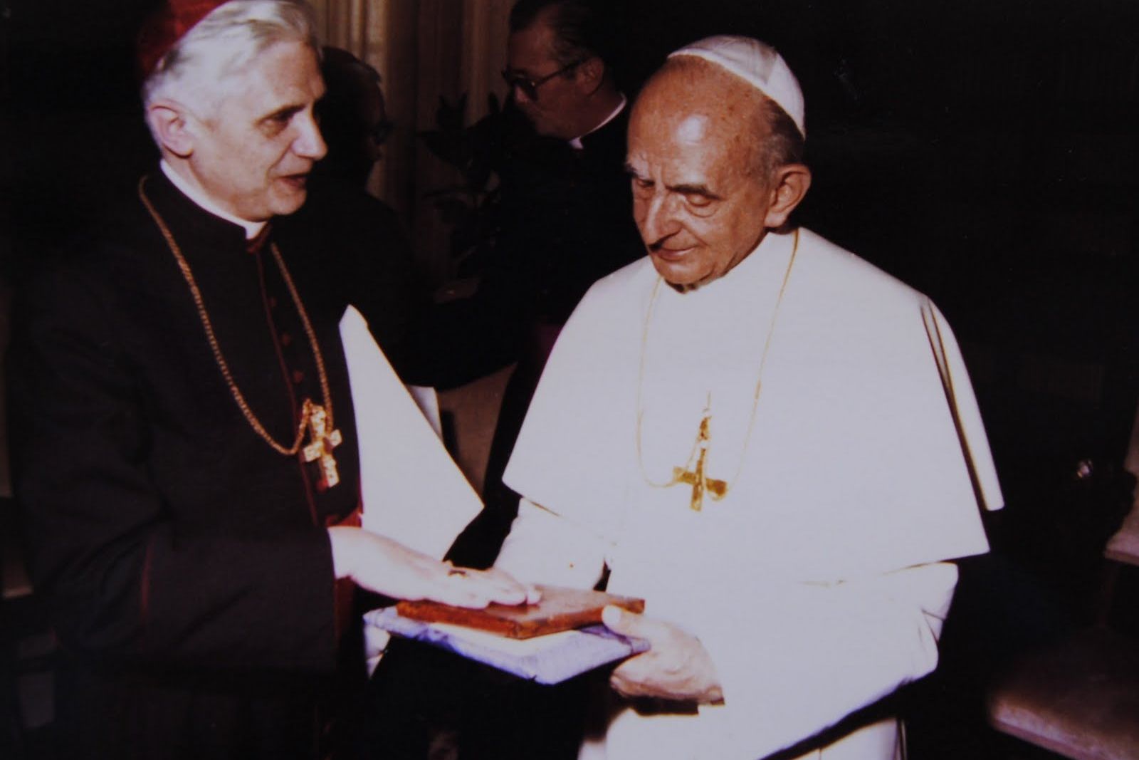 Cardinal Ratzinger in red skullcap presents a book to Pope Paul VI, who stands in white vestments.