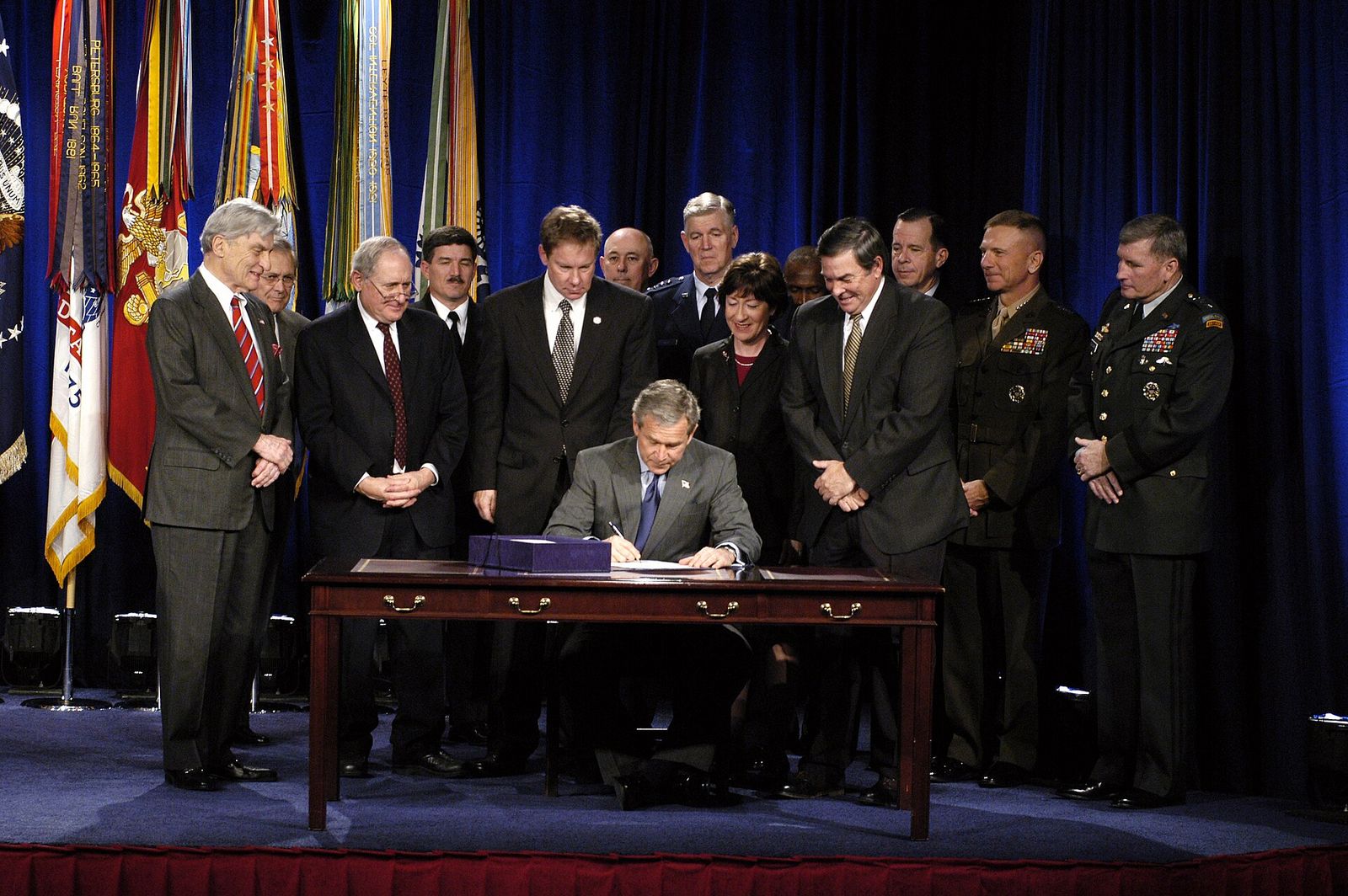 President George W. Bush seated at a desk signing a bill at the Pentagon, surrounded by officials and military officers.