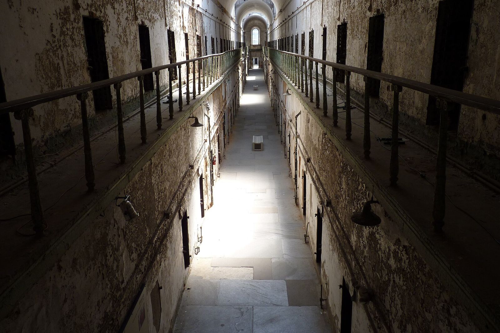 A long, empty, decaying prison cell block with barred walkways and a skylit vault at the far end.