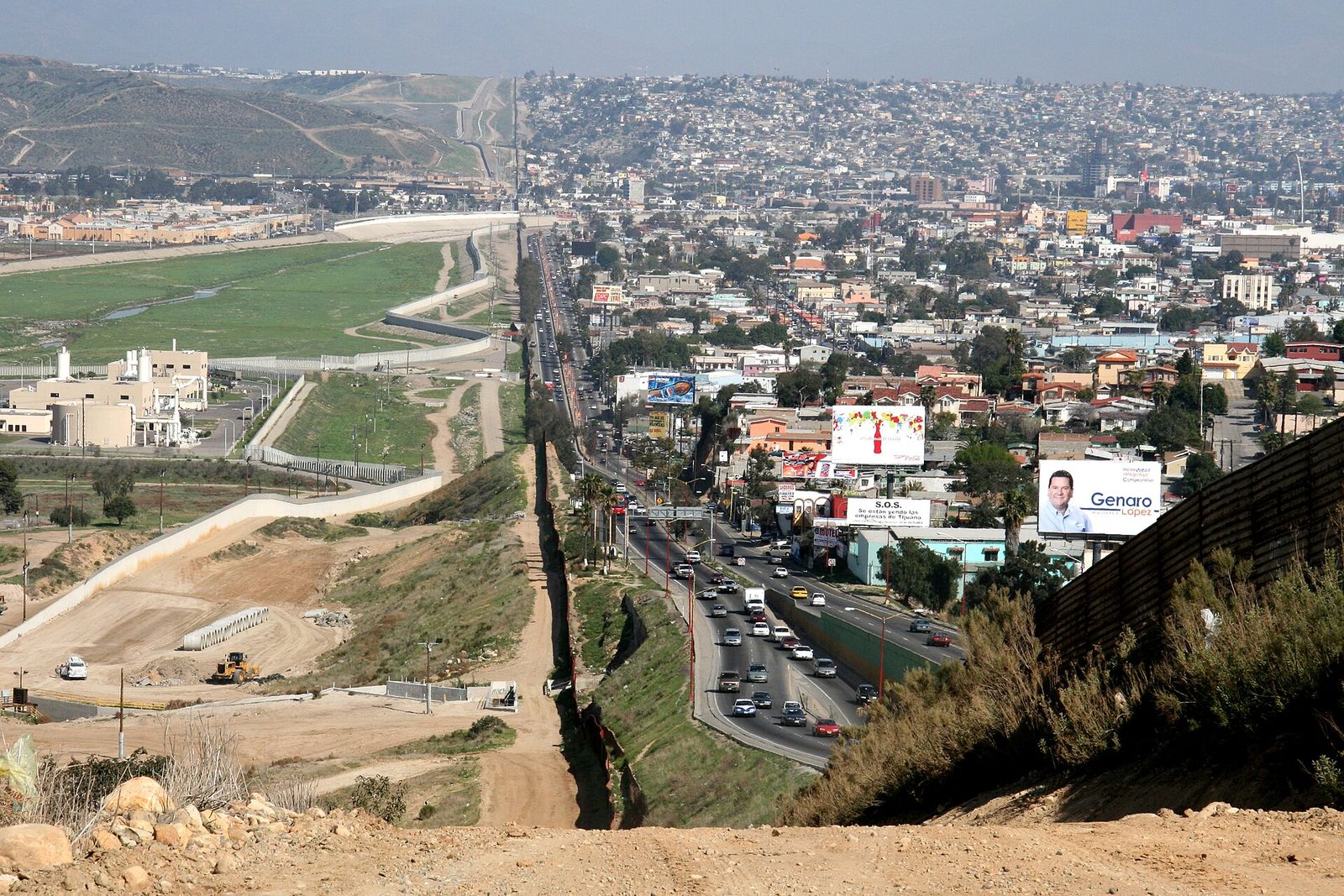 A border fence running down a hillside, with the dense city of Tijuana on one side and open ground on the US side.