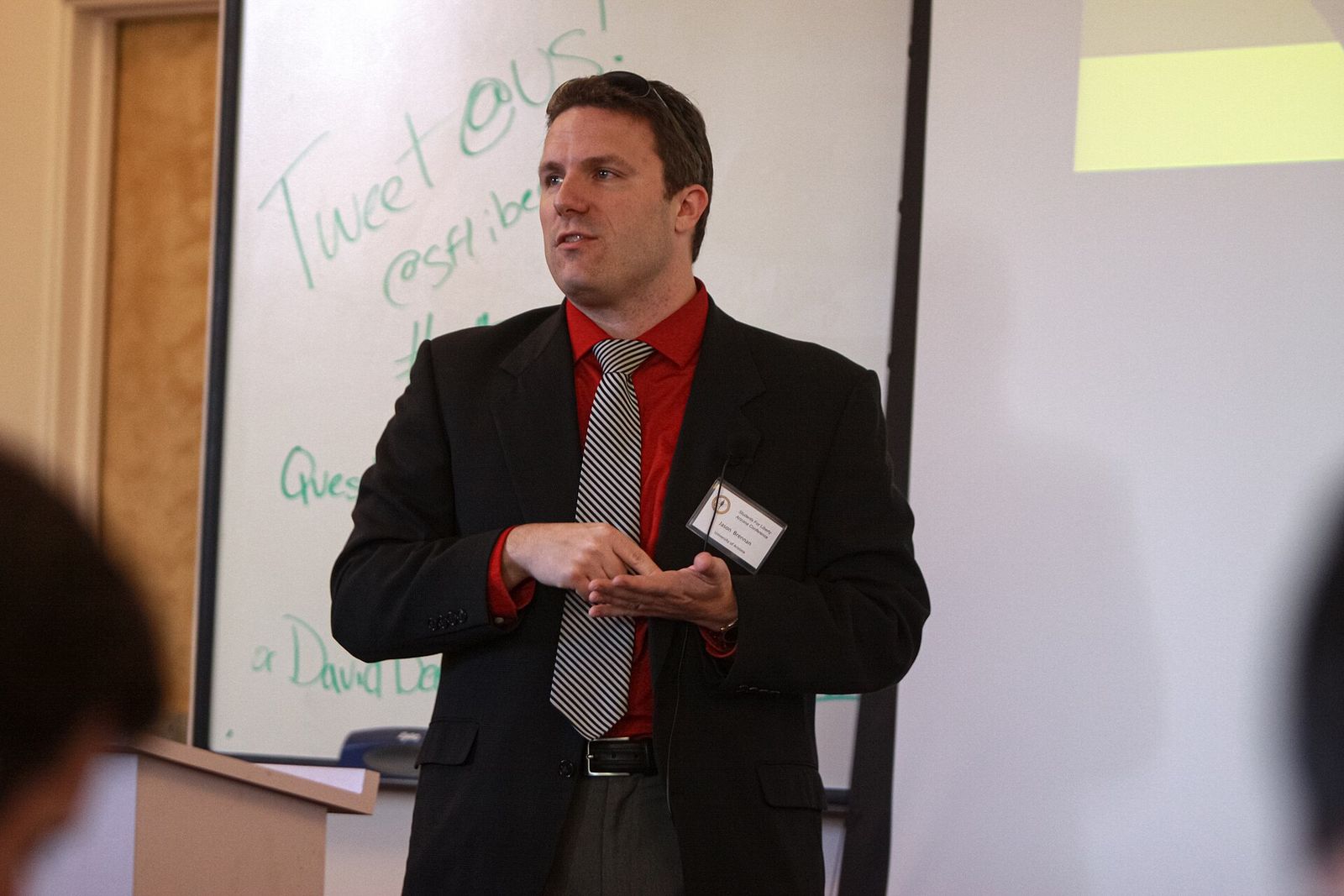 A man in a dark jacket and striped tie speaks beside a lectern in front of a whiteboard.