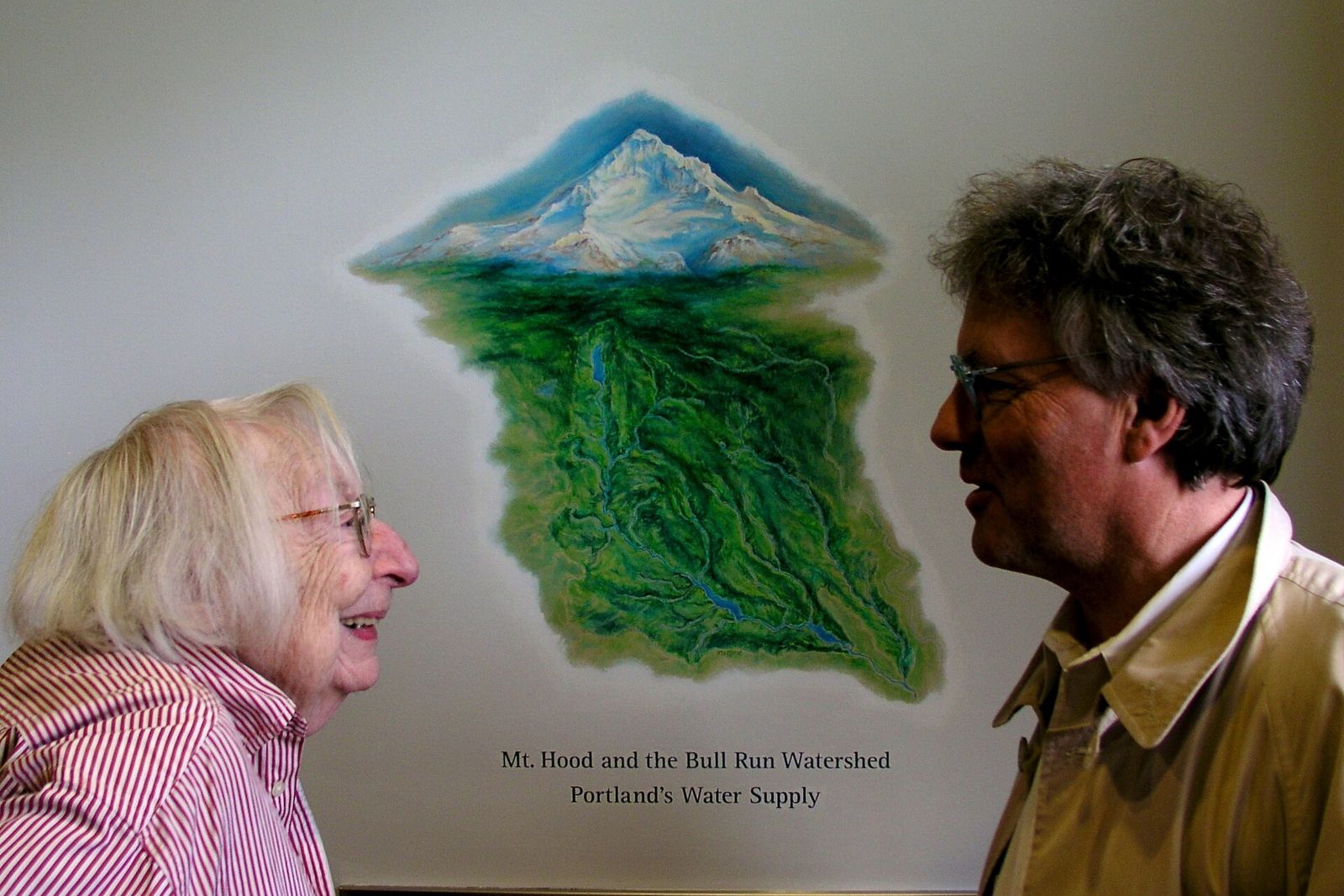 Jane Jacobs, an elderly woman, smiles in conversation with a man in front of a painted map of a watershed.