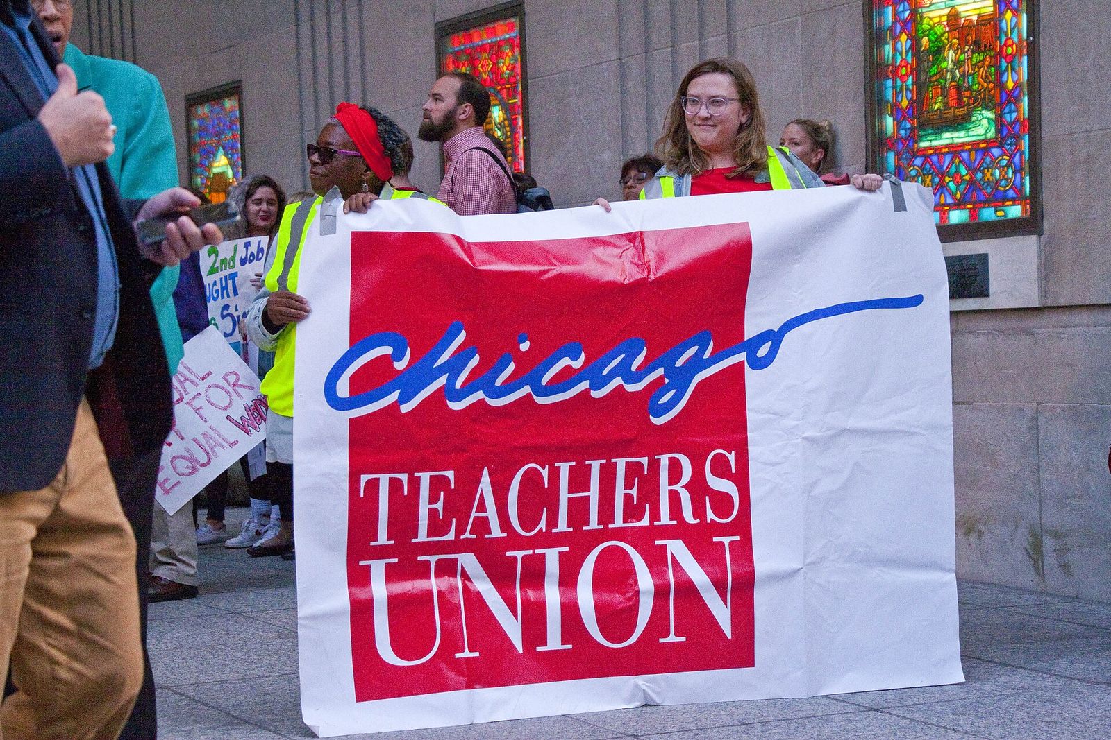 Teachers and allies hold a large Chicago Teachers Union banner outside a building with stained-glass windows.