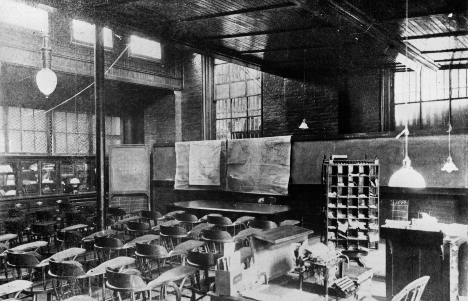 An empty prison classroom with rows of chairs, a blackboard, wall maps and a teacher's desk.