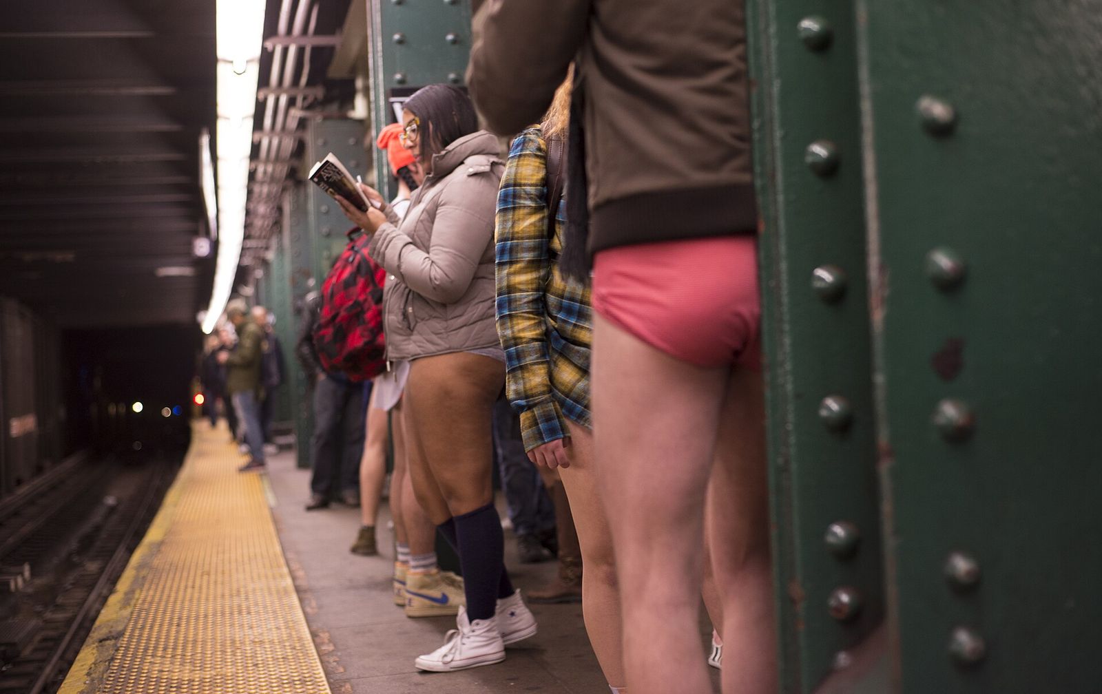 Commuters in coats and bare legs waiting on a New York subway platform beside a green steel column.