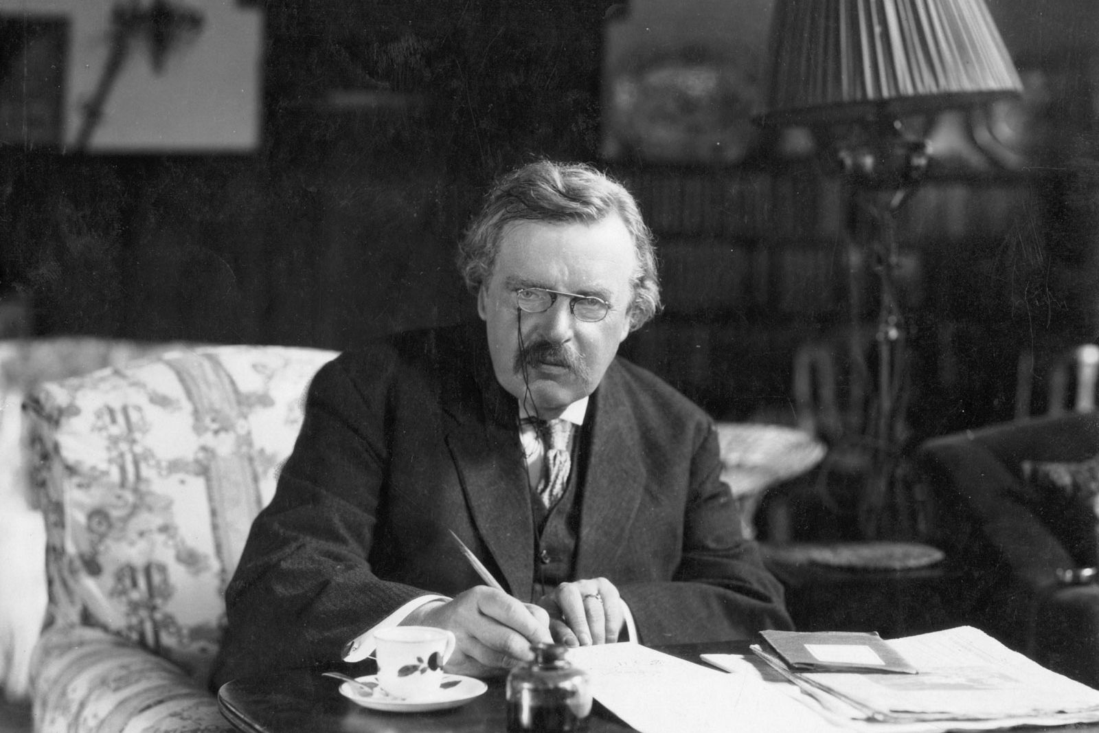 G. K. Chesterton writing at a table, pen in hand, in a book-lined room.