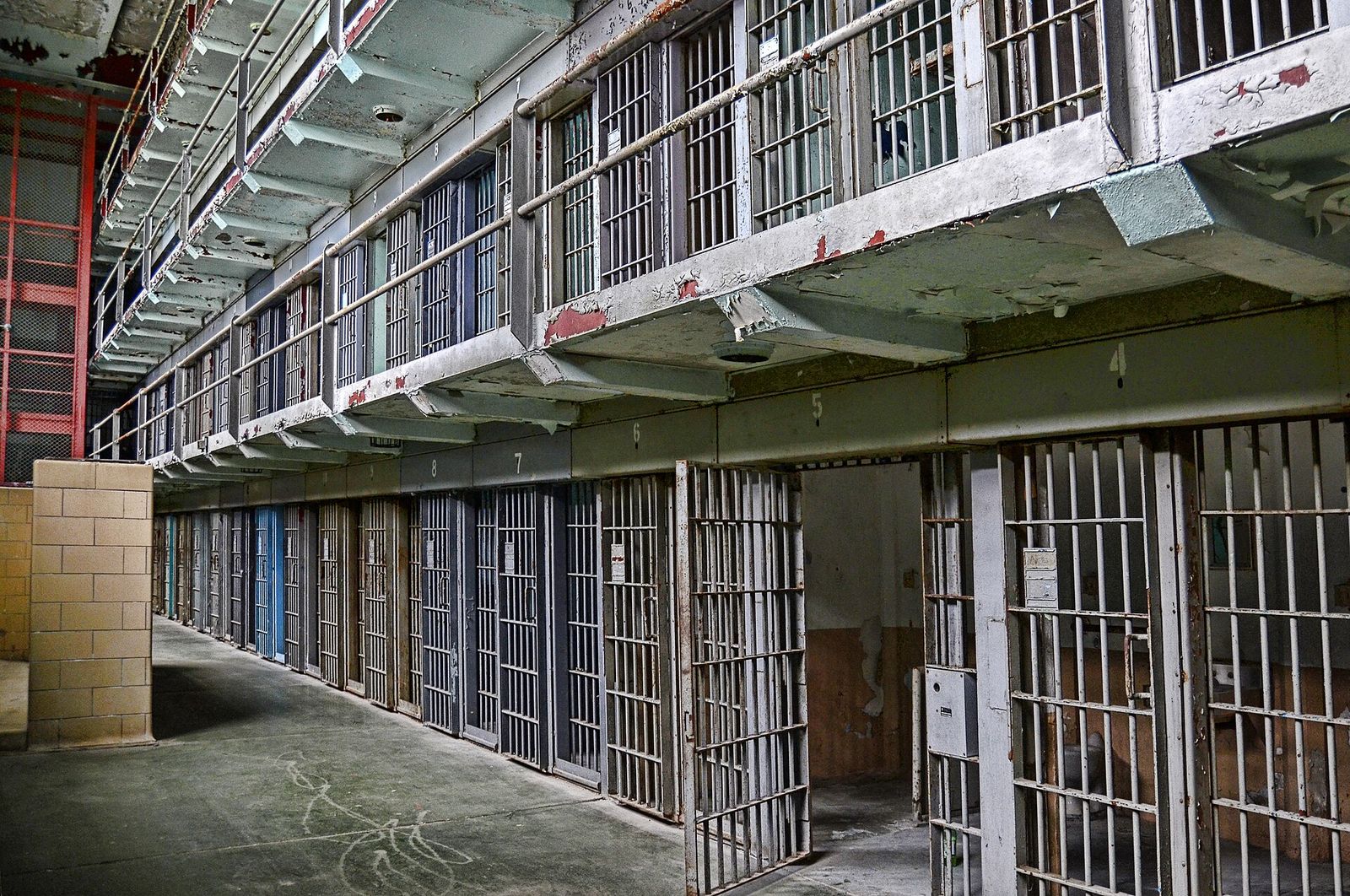 Tiers of barred cells line an older prison cell block with a concrete walkway below.