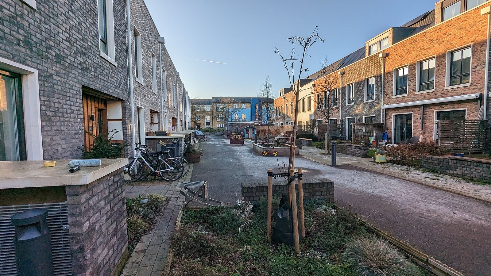 A narrow car-free lane between brick townhouses, with shared planting and bicycles along the edges.