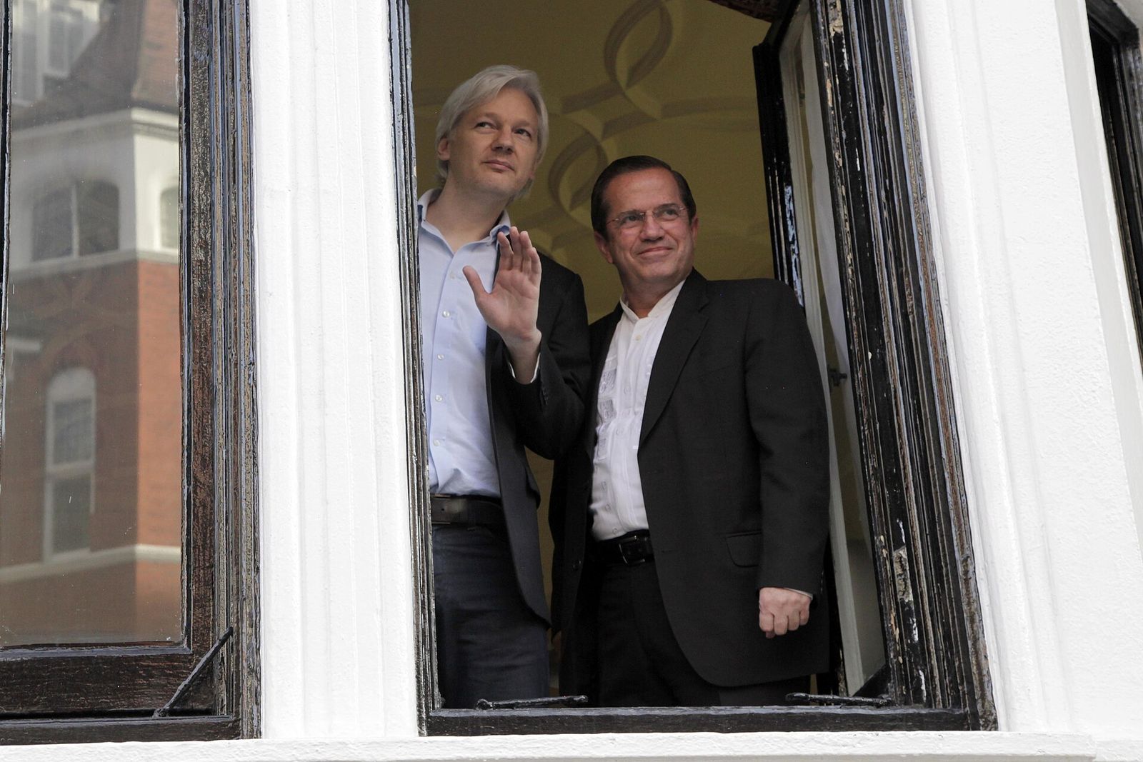 Julian Assange waves from an open window beside Ecuadorian foreign minister Ricardo Patiño.