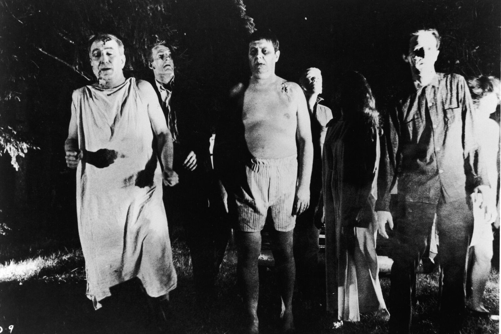 A group of zombies walks through a dark wood in a black-and-white still from Night of the Living Dead.