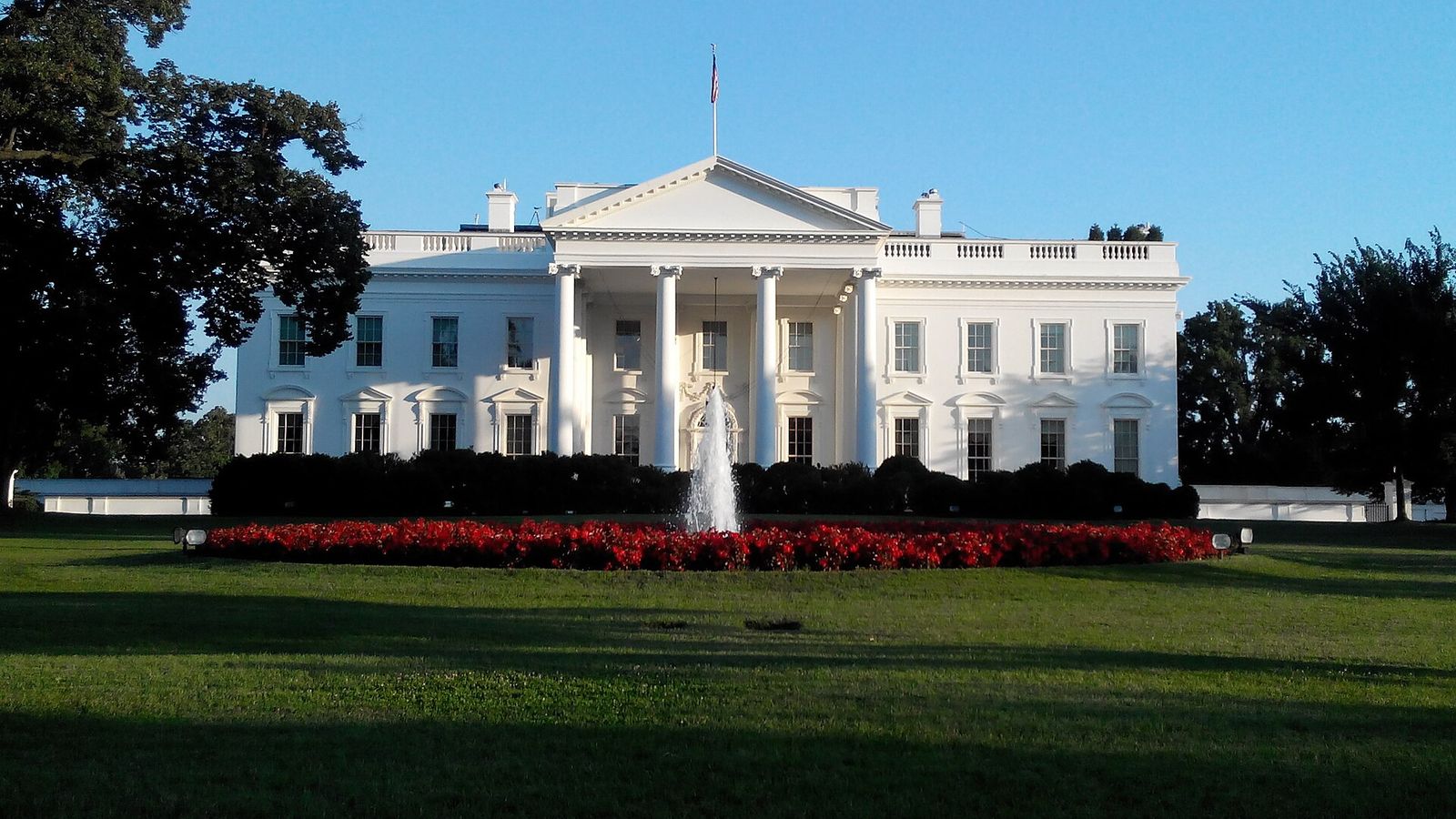 The White House seen across its north lawn, with a fountain and a bed of red flowers in front.