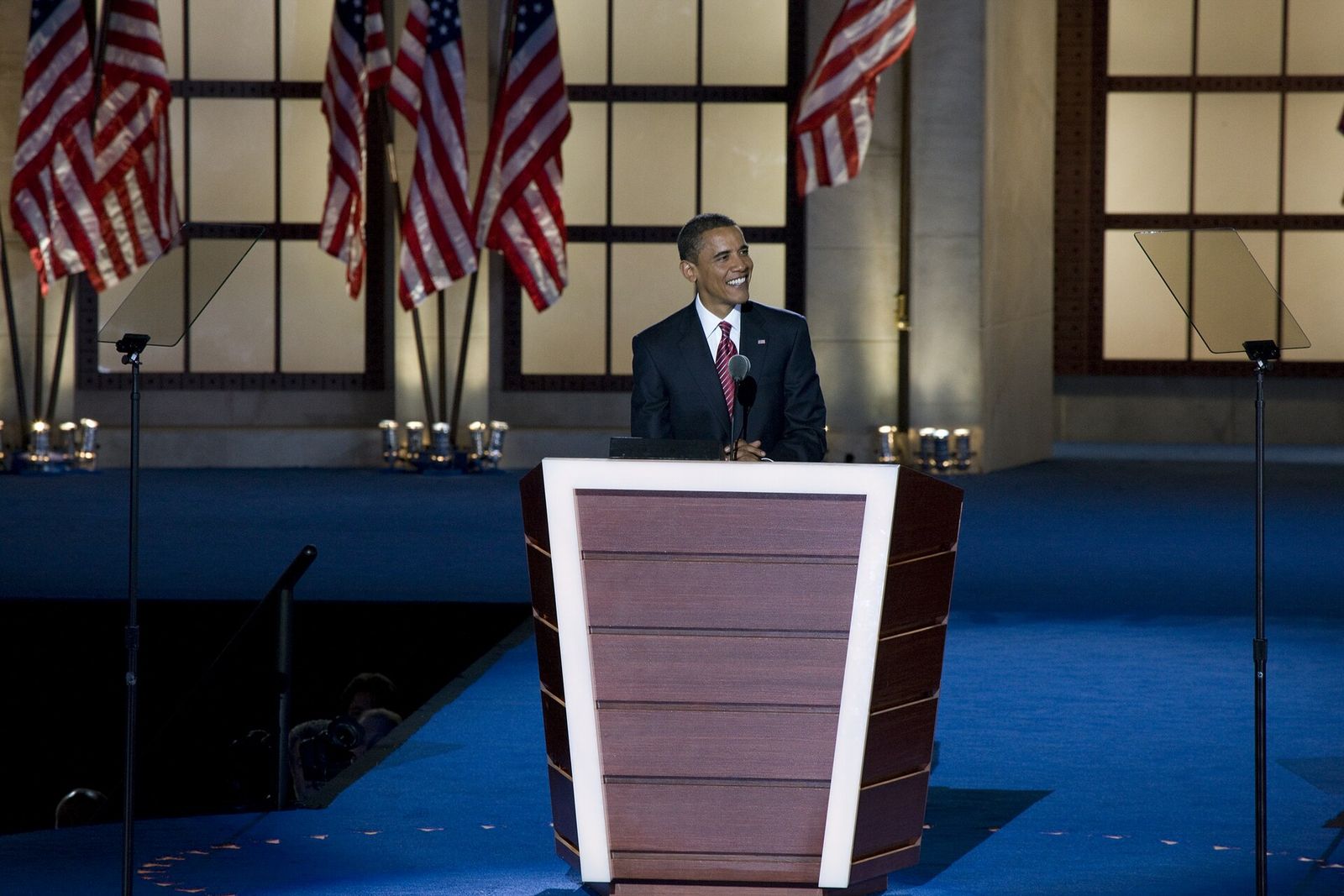 Barack Obama stands smiling at a podium on a blue stage before rows of American flags.