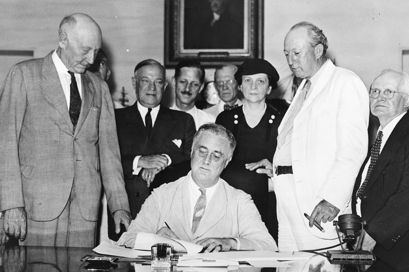 President Roosevelt signing the Social Security Act at a desk, surrounded by legislators and officials.