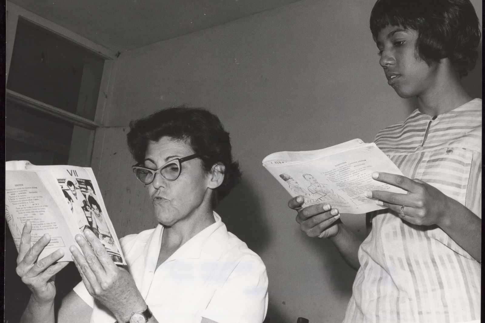 Two women hold open primers and read together in a plain room.