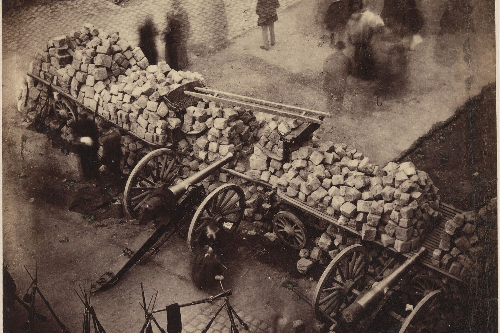 A barricade of stacked paving stones on carts, with cannons and stacked rifles, on a Paris street during the Commune.